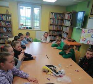 Uczniowie klasy trzeciej siedzą przy dużym stole w gminnej bibliotece. Na stole leżą różne odpadki i śmieci.