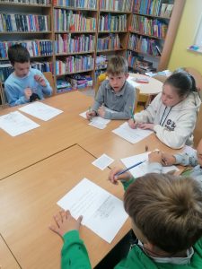Uczniowie klasy trzeciej siedzą przy dużym stole w gminnej bibliotece. Przed nimi leżą kartki papieru a dzieci w dłoniach trzymają ołówki.
