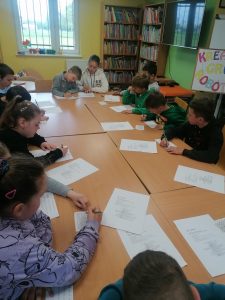 Uczniowie klasy trzeciej siedzą przy dużym stole w gminnej bibliotece. Przed nimi leżą kartki papieru a dzieci trzymają w dłoniach długopisy.