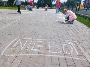Uczniowie na szkolnym boisku rysują kredą domki. Na pierwszym planie widoczny jest narysowany na chodniku napis : NIEBO" wraz ze strzałką skierowaną w górę.