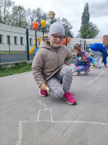 Uczennica kuca na szkolnym boisku. Dziewczynka trzyma w dłoni kredę którą rysuje domek.