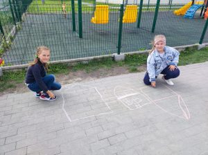 Dwie uczennice kucają na szkolnym boisku. Dziewczynki rysują na chodniku domy.