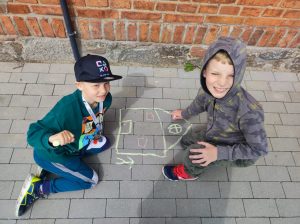 Dwóch uczniów siedzi na chodniku przy szkole. Jeden z nich kredą maluje na chodniku domek.