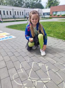Uczennica kuca na chodniku przed budynkiem szkoły. W ręku trzyma kredę. Na chodniku przed nią widnieje rysunek domu.