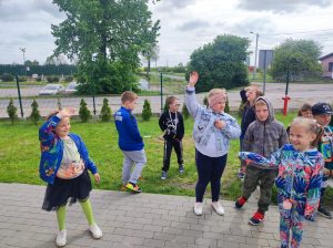 Dzieci stoją na szkolnym boisku. Część z nich podnosi w górę ręce i macha.