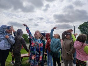 Dzieci stoją na szkolnym boisku. Część z nich podnosi w górę ręce i macha patrząc jednocześnie w niebo.