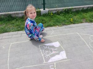 Uczennica kuca na szkolnym boisku. Przed nią widać narysowany kredą dom z kotkiem w oknie.