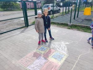 Dwie uczennice pozują do zdjęcia na szkolnym boisku. Przed nimi na chodniku namalowany jest kolorową kredą domek.