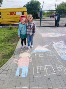 Dwie uczennice pozują do zdjęcia na szkolnym boisku. Przed nimi na chodniku namalowany jest kolorową kredą domek i [postać dziewczynki.