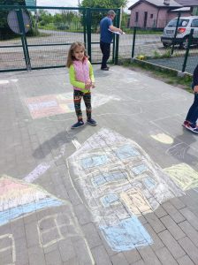 Uczennica stoi na szkolnym boisku. Przed nią narysowany jest kolorową kredą dom.