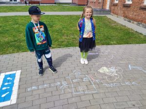 Dwoje uczniów pozuje do zdjęcia na szkolnym boisku. Na chodniku przed nimi narysowany jest domek wraz z dwoma ludzikami podpisanymi Kacper i Amelka.