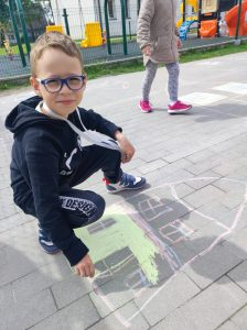 Uczeń kuca na szkolnym boisku. Chłopiec rysuje na chodniku przed sobą dom.
