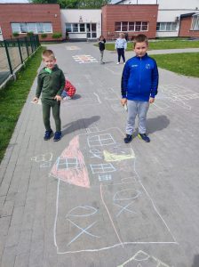 Dwóch chłopców stoi na szkolnym boisku. Przed nimi na chodniku narysowany jest kredą dom.