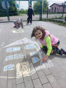 Dziewczynka pochyla się nad narysowanym przez siebie na szkolnym boisku domem. W tle widać innych pracujących uczniów.