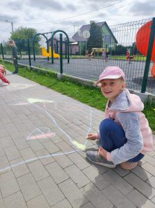 Uczennica kuca na szkolnym boisku. Dziewczynka trzyma w dłoni kredę którą rysuje domek.