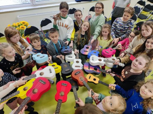 Uczniowie biorący udział w zajęciach ustawieni są w okrąg i wyciągają przed siebie swoje instrumenty. Ukulele stykają się ze sobą pudłami rezonansowymi.