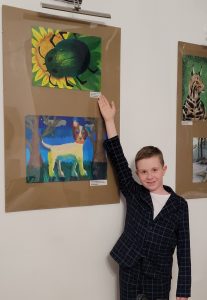 Wyróżniony uczeń stoi przy zawieszonej na ścianie galerii nagrodzonych prac. Chłopiec wysoko podnosi w górę prawą dłoń, którą wskazuje swoją pracę.