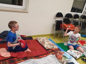 Dwóch chłopców siedzi na podłodze wśród koców. Chłopcy rzucają do siebie piłeczkę, która przyczepia się do trzymanych przez nich paletek. Jeden z chłopców trzyma w dłoni piłeczkę i planuje nią rzucić do kolegi.