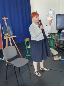 Nauczycielka prezentuje wykonaną przez siebie w stylu decoupage deskę do krojenia. Nauczycielka podnosi w górę deskę trzymaną w jednej z dłoni. W drugiej trzyma mikrofon.