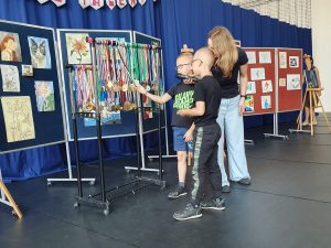 Dwóch uczniów wraz z nauczycielką stoi przy stelażu z medali. Jeden z nich trzyma w dłoni wiszący medal. Nauczycielka pochyla się nad chłopcami i trzyma przy buzi jednego z nich mikrofon.