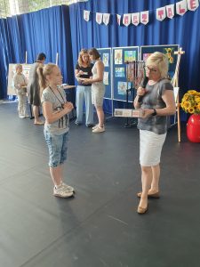 Pani Dyrektor wręcza nagrodę uczennicy, która stoi naprzeciwko niej. Pani Dyrektor przygląda sie trzymanemu w dłoniach pudełku.