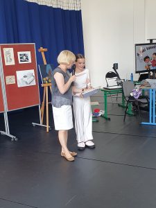 Pani Dyrektor wręcza nagrodę uczennicy za zwycięstwo w konkursie. Obie stoją obok siebie na środku sali. Pani Dyrektor trzyma mikrofon a uczennica ubrana jest na biało.