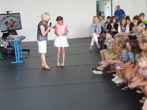 Pani Dyrektor wręcza nagrodę stojącej obok uczennicy. Dziewczynka przygląda się otrzymanej książce. Obok widać siedzących na widowni innych uczniów.