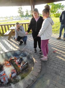 Dwie uczennice pieką kiełbaski w ognisku. Obok nich kuca jeden z rodziców.