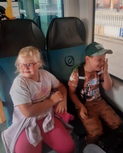 Dwoje uczniów siedzi obok siebie w pociągu. Chłopiec się smieje i przeciera palcem oko. Dziewczynka patrzy w bok.