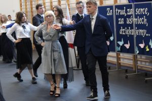 Zdjęcie wykonane podczas poloneza. Na pierwszym planie widoczna jest Pani Dyrektor z uczniem. Oboje mają złączone dłonie wyciągnięte przed siebie. W tle widać kolejne tańczące pary.