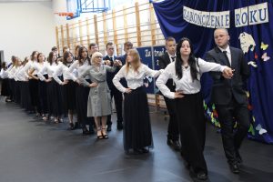Uczniowie klasy ósmej stoją w parach jedna za drugą. W jednej z par stoi również Pani Dyrektor. Dziewczyny ubrane są w białe koszule i długie czarne spódnice a chłopcy w garnitury. Pierwsza para ma złączone i wyciągnięte przed siebie dłonie.