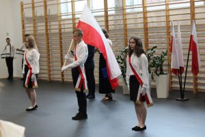 Troje uczniów stoi w jednym rzędzie w równych odległościach od siebie. Chłopiec stojący po środku trzyma flagę Polski. Wszyscy mają przełożone przez ramię biało-czerwone szarfy.