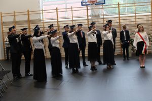 Uczniowie klasy ósmej podczas ślubowanie. Ustawieni są w dwóch rzędach z wyciągniętymi przed siebie rękoma. Na głowach mają birety. Obok stoi uczennica z pocztu flagowego szkoły z biało-czerwoną szarfą przełożoną przez ramię.
