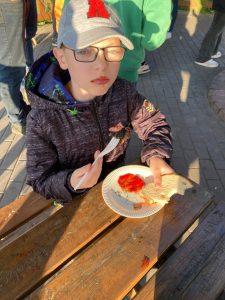 Uczeń siedzi przy stoliku i je kiełbaskę, którą trzyma na widelcu. Chłopiec ma czerwone usta od ketchupu. Przed nim na stoliku leży talerzyk z plama ketchupu o który chłopiec opiera rękę z kawałkiem chleba.