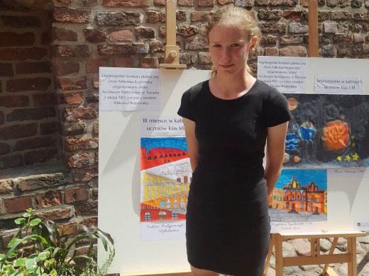 Zdjęcie uczennicy Natalii Matyjaszczyk pozującej na tle sztalugi z jej nagrodzoną pracą. Dziewczynka ubrana jest w czarną krótką sukienkę.