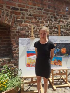 Zdjęcie uczennicy Natalii Matyjaszczyk pozującej na tle sztalugi z jej nagrodzoną pracą. Dziewczynka ubrana jest w czarną krótką sukienkę.