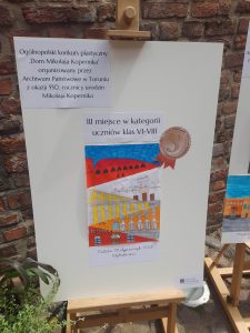 Zdjęcie naklejonej na dużej kartce białego papieru i umieszczonej na sztaludze pracy uczennicy. Nad Pracą napis "III miejsce w kategorii uczniowie klas VI - VIII" a w prawym górnym rogu przyklejona karteczka z informacją "Ogólnopolski konkurs plastyczny "Dom Mikołaja Kopernika" organizowany przez Archiwum Państwowe w Toruniu z okazji 550 rocznicy urodzin Mikołaja Kopernika".