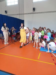 Uczniowie tworzą węża idąc za prowadzeni przez Panią w żółtej sukience. Wąż przechodzi obok muzyków orkiestry.