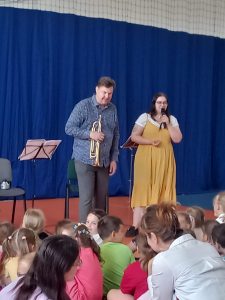 Dwoje muzyków stoi przed uczniami. pan trzyma w dłoni trąbkę a Pani mikrofon.