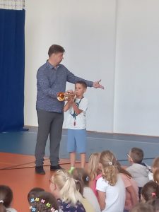 Pan stoi przy uczniu, który gra na trąbce. mężczyzna patrzy w bok i ma wyciągniętą lewą rękę przed siebie.