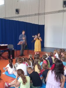 Dwoje muzyków z Toruńskiej Orkiestry Symfonicznej stoi przed siedzącymi na podłodze uczniami. Oboje trzymają w dłoniach instrumenty muzyczne.