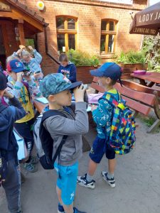 Uczniowie stoją przed jednym z budynków osady leśnej Barbarka. Część dzieci spogląda przez trzymane w dłoniach lornetki.