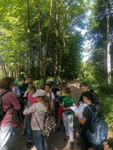 Uczniowie stoją w lesie. Część z nich spogląda na trzymane w dłoniach kartki.