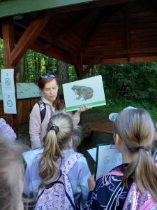 Uczniowie stoją na przeciwko Pani, która trzyma w dłoni kartkę z rysunkiem żaby.