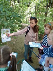 Dzieci trzymają podkładki z kartkami w dłoniach i patrzą na rysunek, który wskazuje im Pani. Rysunek przyczepiony jest do drzewa i przedstawia jego grafikę.
