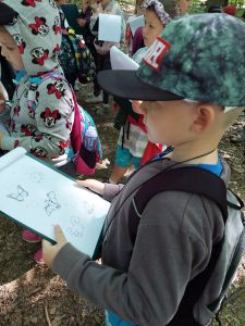 Chłopiec trzyma w dłoni podkładkę z kartką na której widać rysunki zwierząt. W tle widać stojące inne dzieci.