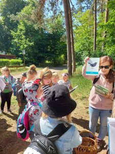 Uczniowie stoją na polanie zgromadzeni wokół Pani. Kobieta trzyma w wyciągniętej w górę ręce kartkę z rysunkiem ptaka.