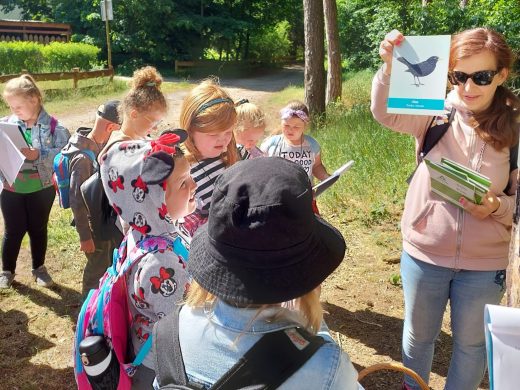 Uczniowie stoją na polanie zgromadzeni wokół Pani. Kobieta trzyma w wyciągniętej w górę ręce kartkę z rysunkiem ptaka.