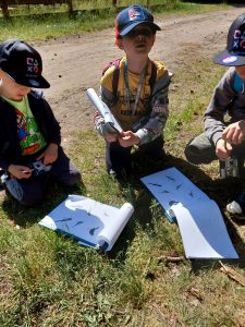Troje chłopców kuca na trawie. Przed nimi leżą kartki z rysunkami ptaków.