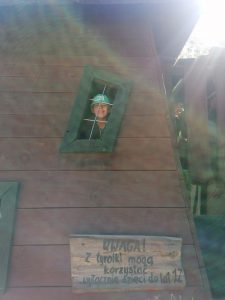 Chłopiec wychyla sie przez okienko w drewnianym budynku. Pod okienkiem na drewnianej desce wyryty jest napis: "UWAGA! z tyrolki mogą korzystać wyłącznie dzieci do lat 12".